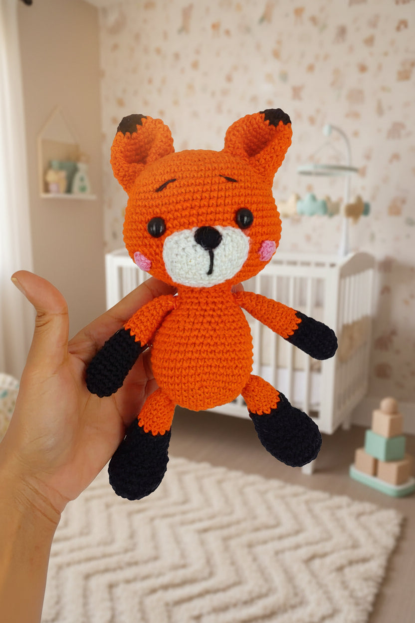 A crocheted orange fox plush toy with black paws and feet, held in a person's hand against a lush green forest background.
