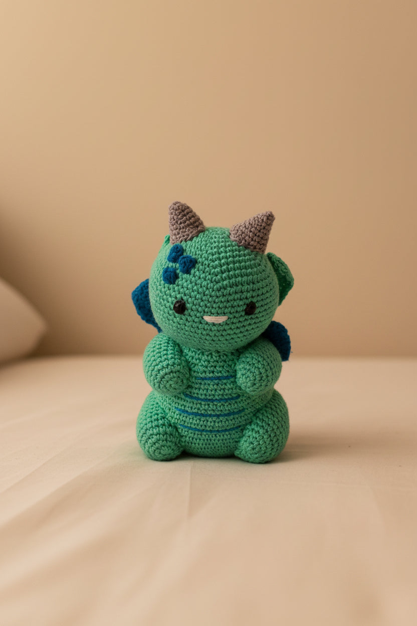 A small crocheted dragon toy in bright green with blue accents, blue wings, and brown horns, sitting against a plain light gray background.
